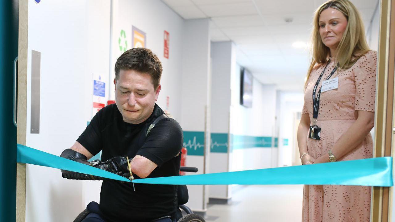 Alex Lewis cuts a blue ribbon with a pair of scissors. A woman wearing pink and can be seen looking on in the background