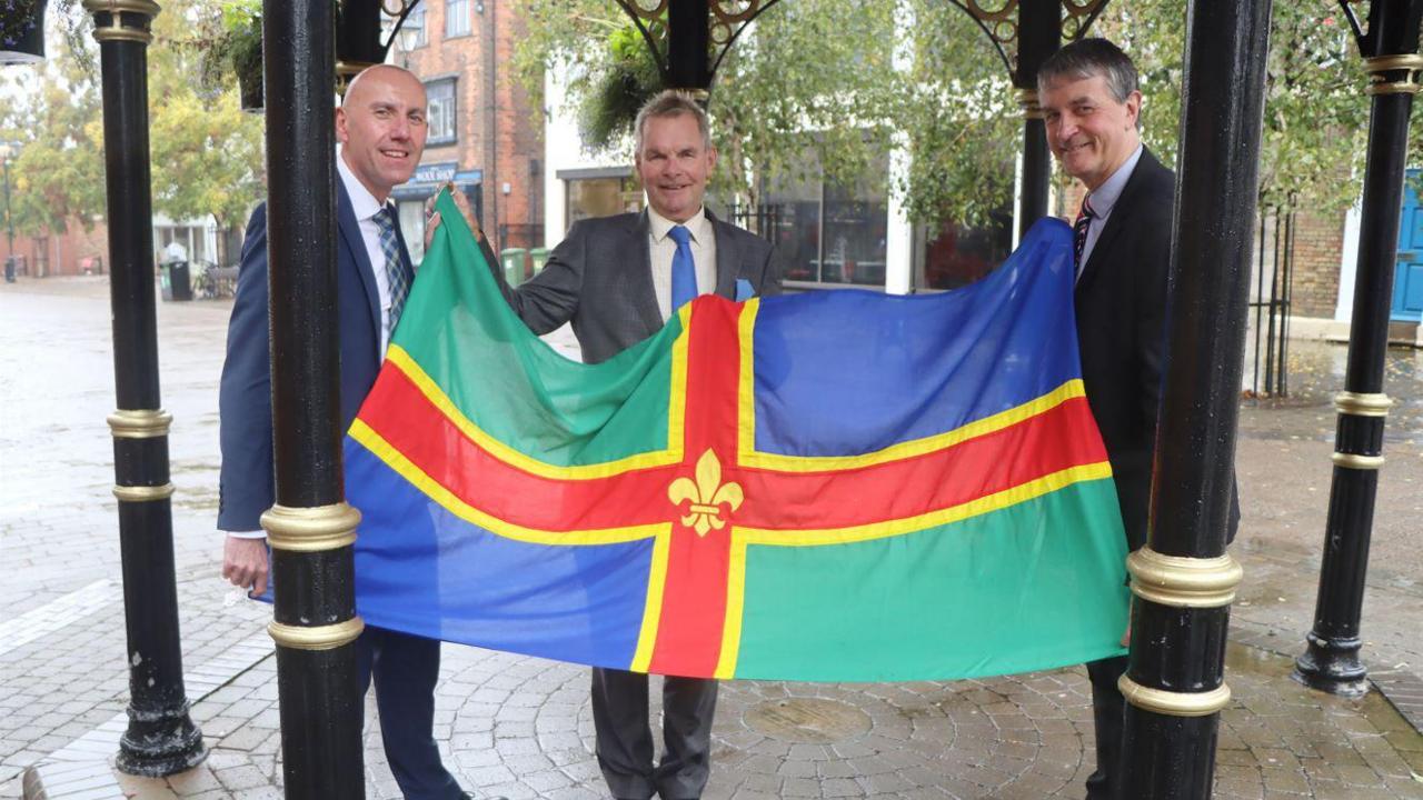 Council Leader for North East Lincolnshire, Councillor Philip Jackson (right) with Councillor Rob Waltham MBE, the Leader of North Lincolnshire Council (left) and the Leader of Lincolnshire County Council, Councillor Martin Hill OBE pictured on Lincolnshire Day 2024. They are all wearing suits and are holding a flag. 