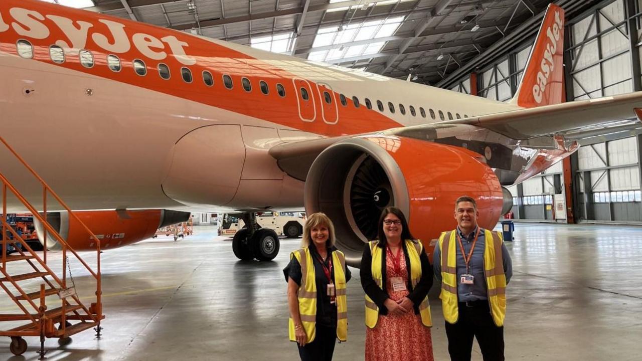 Rachel stands in a hangar in front of an EasyJet plane with its trademark orange and white colours. She is flanked by two people and they all wear high vis yellow vests and red lanyards.