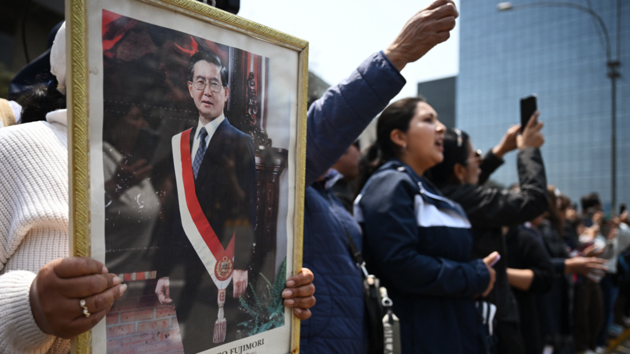 Supporters of the late Alberto Fujimori hold a framed image of the former president who is draped in a sash in the colours of Peru's national flag. To the right more people wave their hands in the air as they wait for Fujimori's hearse to pass the crowd