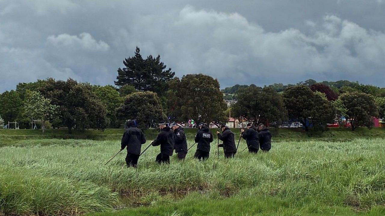Seven police officers are seen in the middle of a field of tall grass with a body of water to the left and trees in the background beneath a moody cloudy sky. 