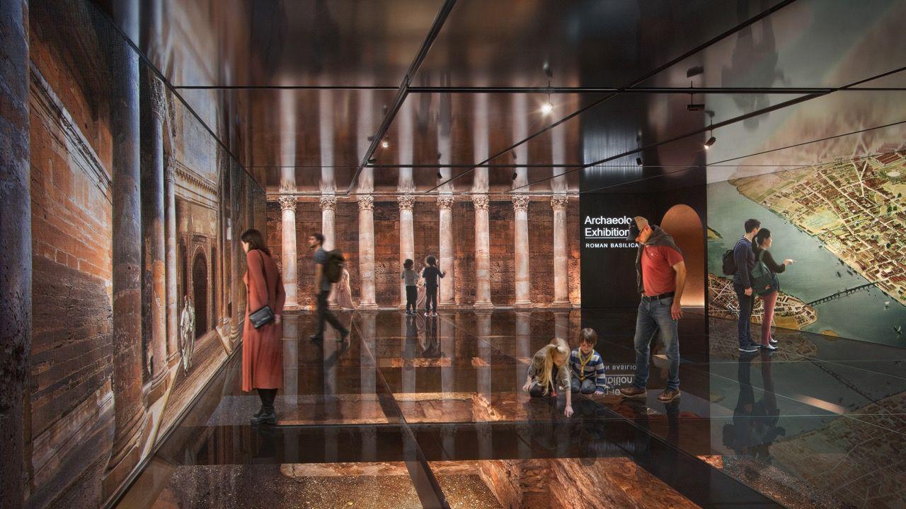 An artist's impression showing what the exhibition might look like in the finished building. It is a dimly lit large room with a glass floor looking down onto the exposed Roman walls and artworks on each wall showing what the building would have looked like with large columns at its entrance. 