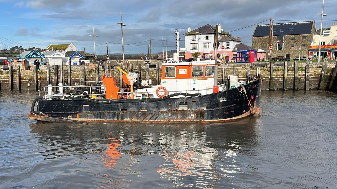 Side view of dredging vessel Doonhammer in Bridport harbour. The boat is about 15 metres long and has a black hull with an orange and white cabin. In the background is the stone harbour wall lined with wooden mooring posts. Along the quayside are lines of wooden takeaway huts and some Victorian-era buildings, including a brick warehouse, a white and pink painted pub and some terraced houses and shops.