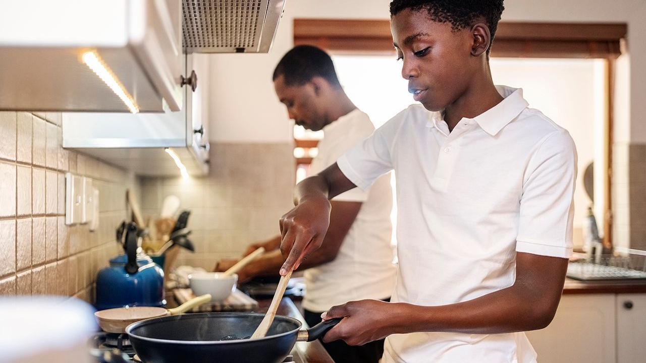 Stock photo showing a teenager using a frying pan while cooking in the kitchen with his father in the background