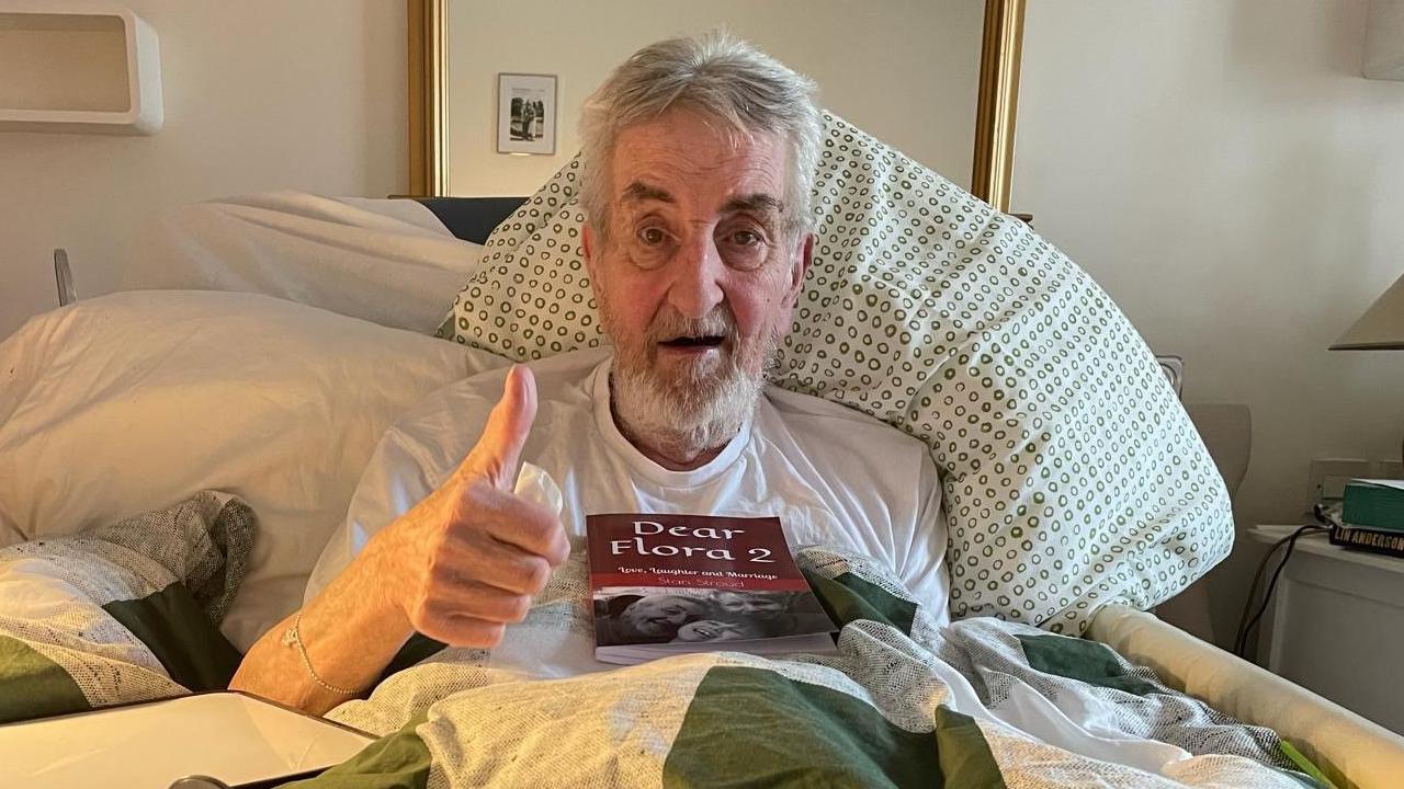 A man with short grey hair and a beard, is wearing a white T-shirt. He is sitting up in a bed, with a pillow behind him, and is looking at the camera and giving a thumb's up with his right hand. On his chest is a book called "Dear Flora 2".