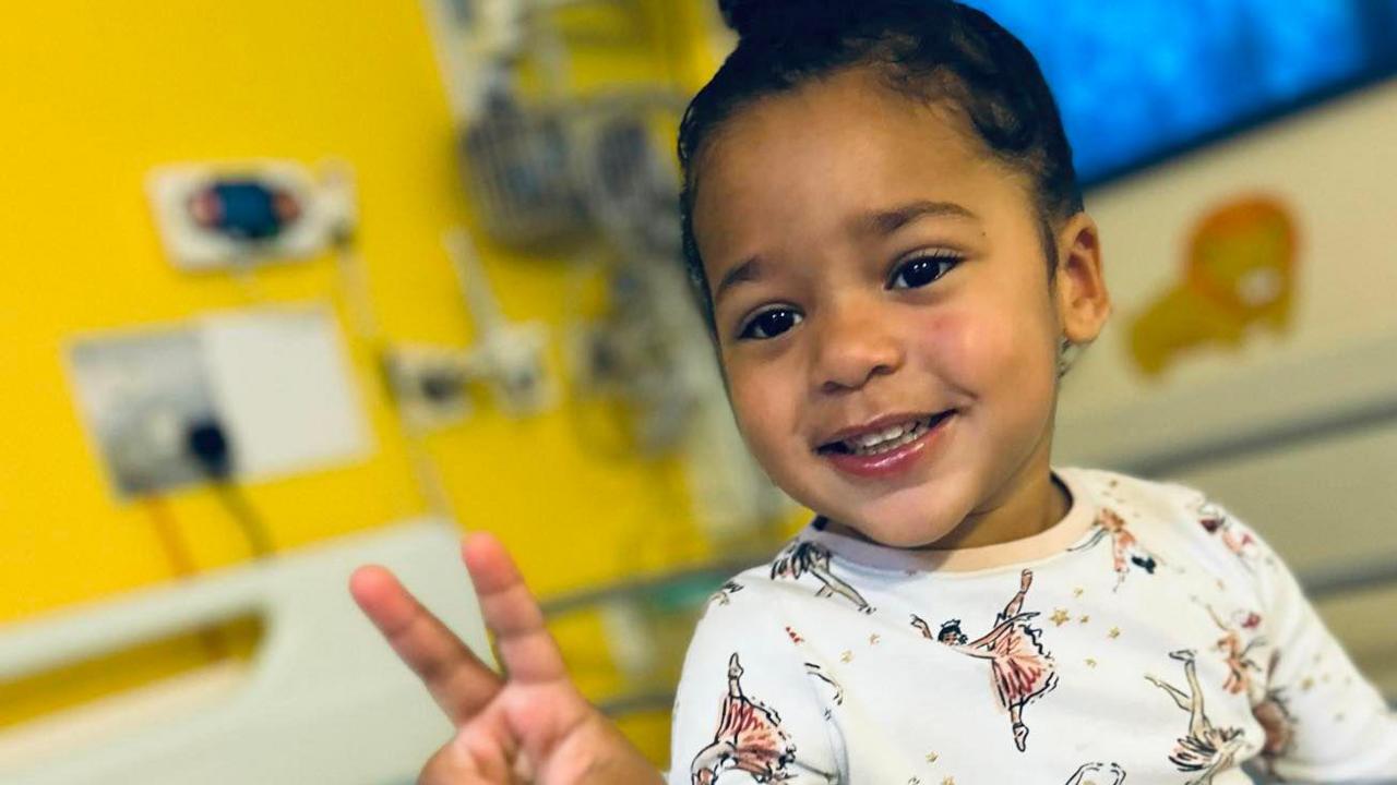 A young girl sitting in a hospital bed smiling and doing a peace sign