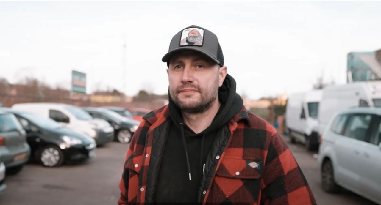 A man in a car park walking towards the camera. He is wearing a red lumberjack shirt over a black hoodie, and a baseball cap. He has a goatee beard and a moustache.