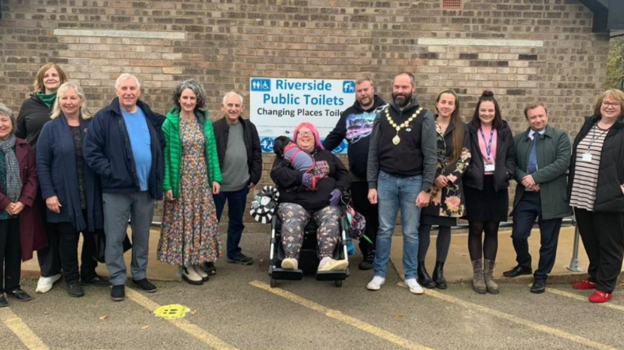 A group of people lined up by a wall with a sign "Riverside Public Toilets".