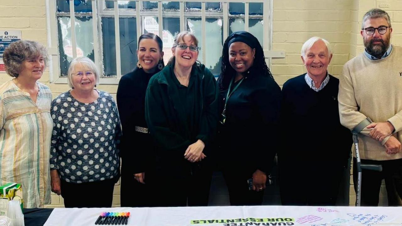 A group of food bank volunteers, staff and supporters pose for a group photo next to a table. There are seven people including Michelle, the Operations Manager, who is at the centre of the photo and wears a green foodbank lanyard.