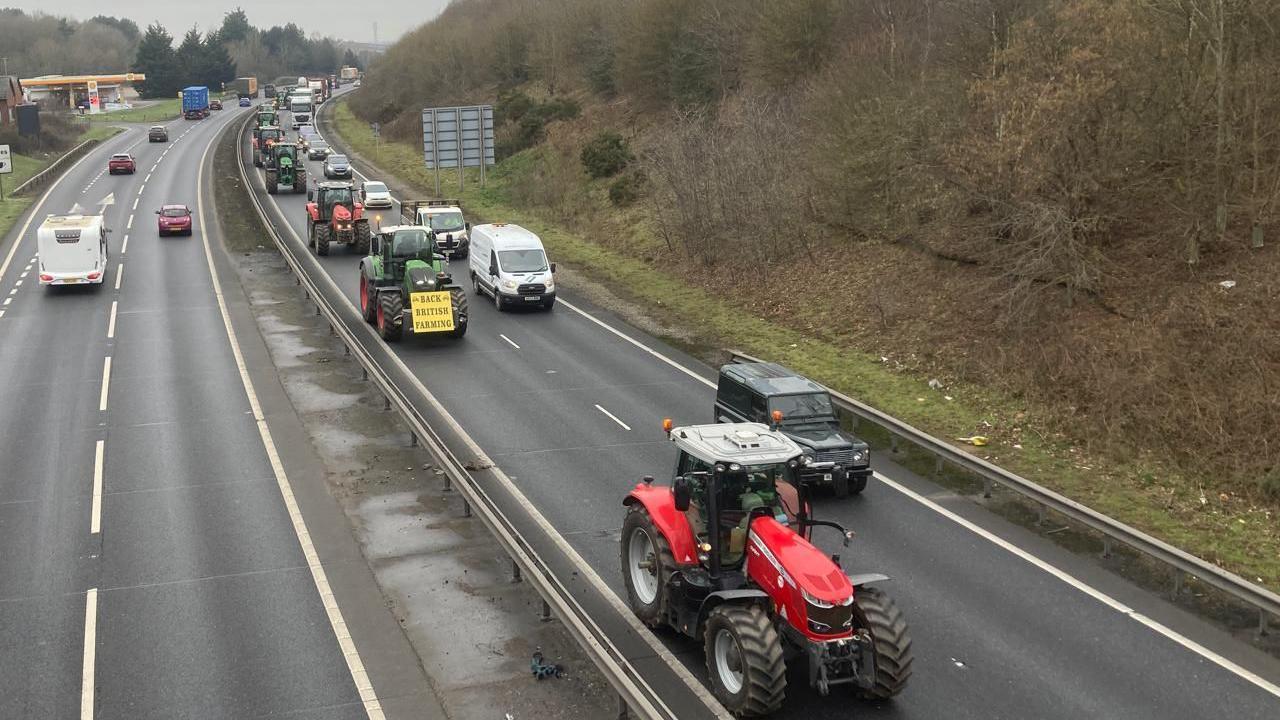 Tractors are pictured from above heading down one lane of the A14. They drive toward the camera while other vehicles drive past them in the parallel lane. 