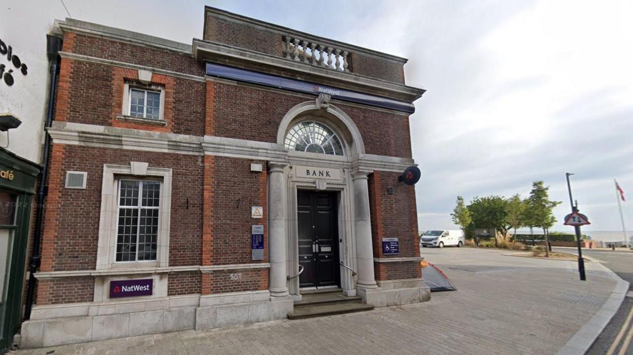 The distinctive former NatWest bank - a dark-brick building with decorative stonework. A black door is surrounded by a tall arched doorway, with stone columns to either side and the word "bank" carved in capital letters on a stone lintel. The sea can be seen in the distance.