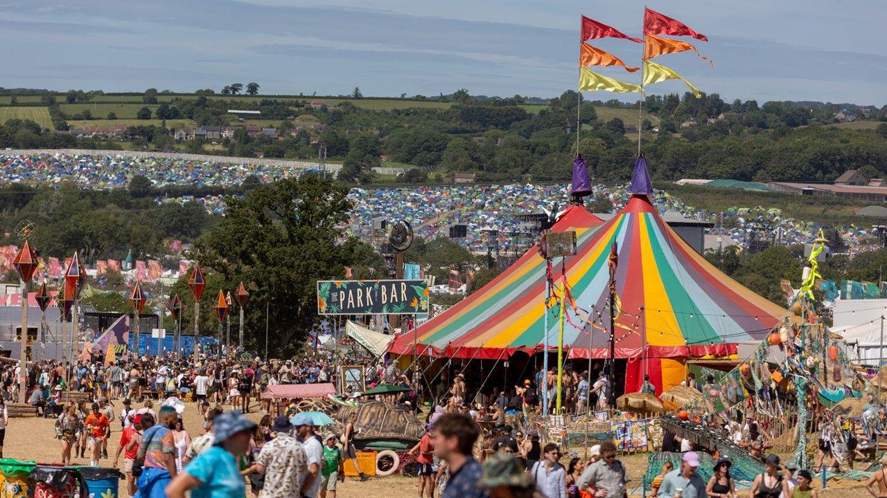 A multi-coloured big top tent surrounded by people and a view across fields of tents at Glastonbury