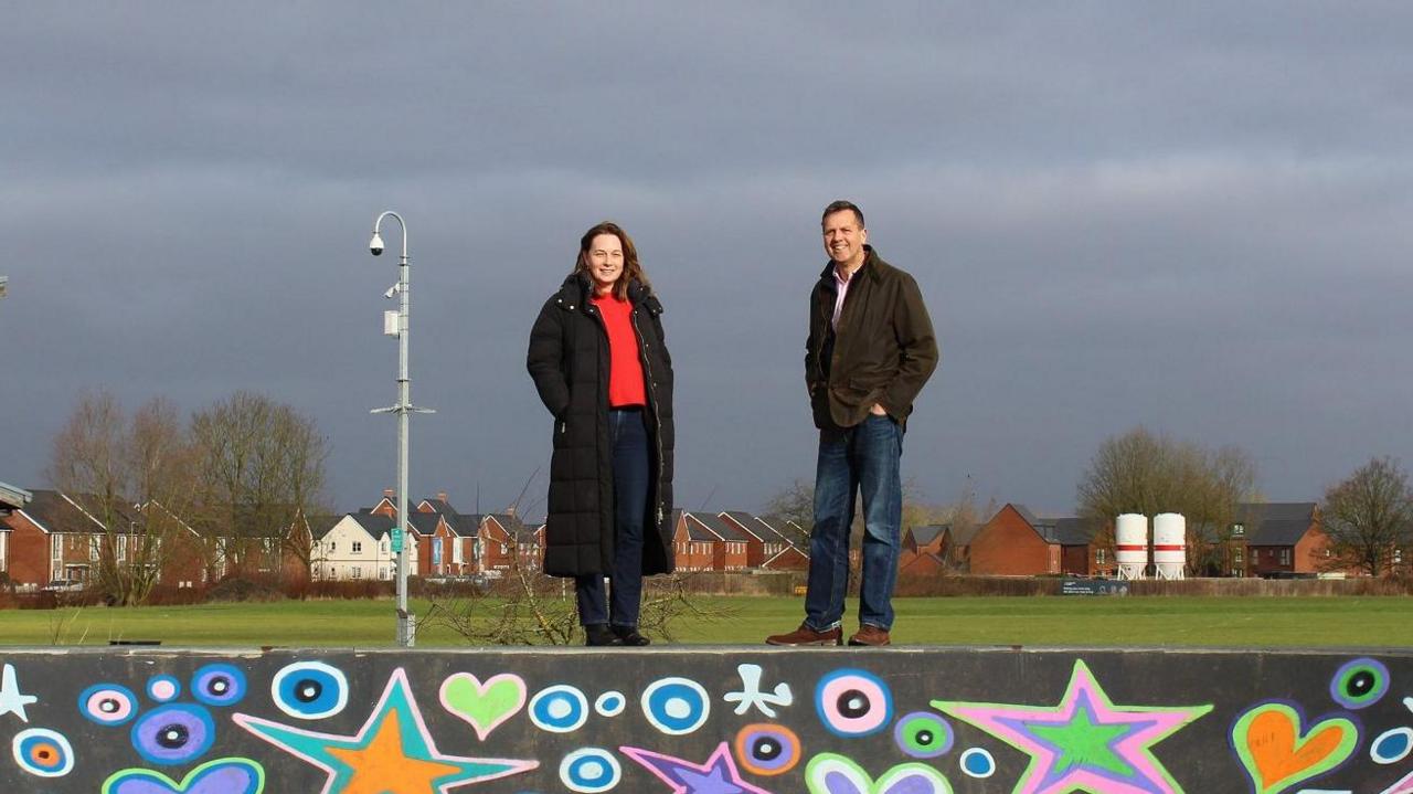 A woman and a man are stood on top of a colourful mural with colourful stars and circles. There is a pole and a CCTV camera visible nearby.