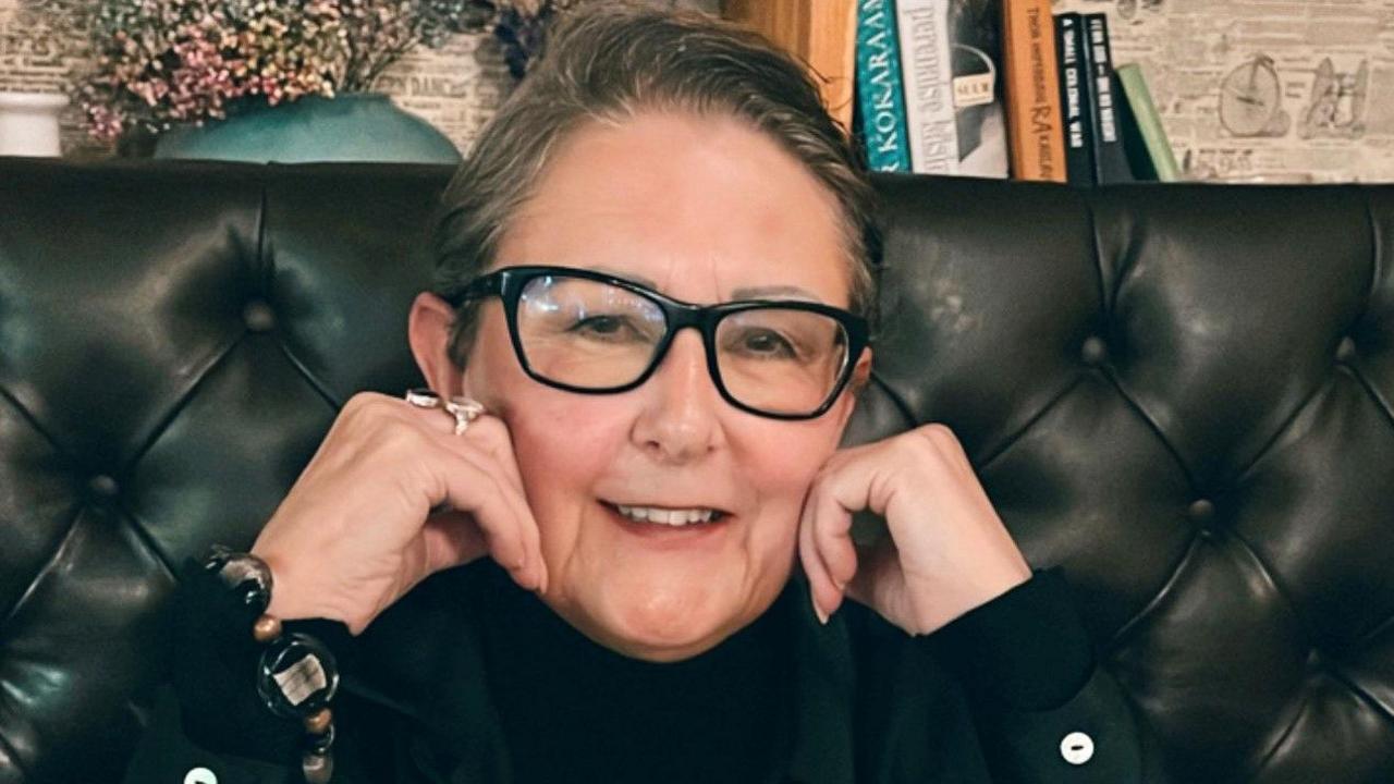 A close-up of Ms Danahay with her hands either side of her face. She has dark slicked back hair and is wearing black rimmed glasses. She is sitting in a restaurant.