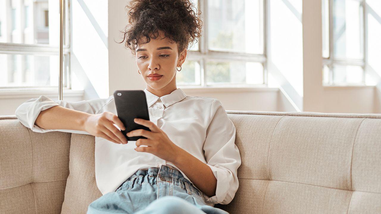 A woman wearing a white shirt and jeans sits on a sofa looking at a phone