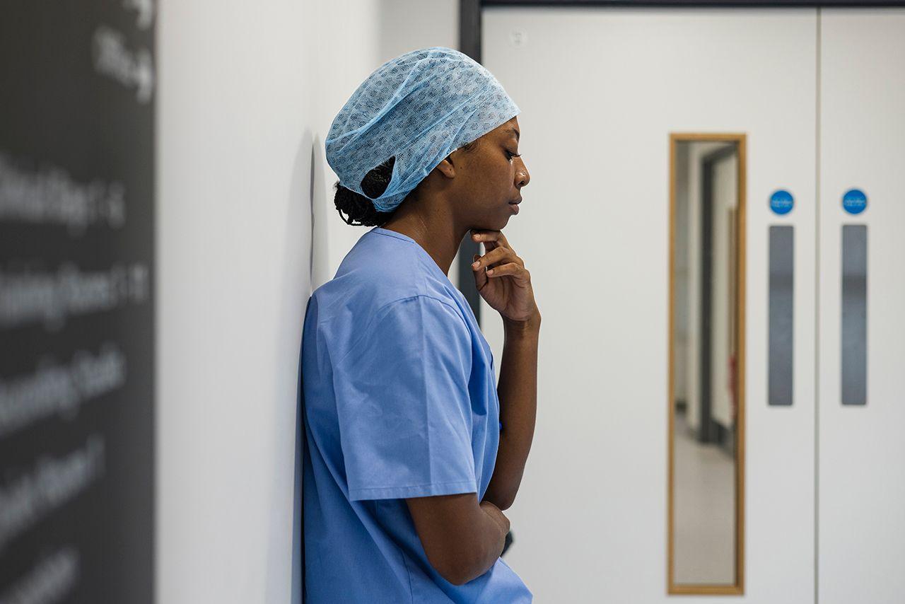 An NHS hospital worker wearing scrubs and a hair net stands against a wall inside a hospital in Newcastle while looking pensive. Stock photo illustration.