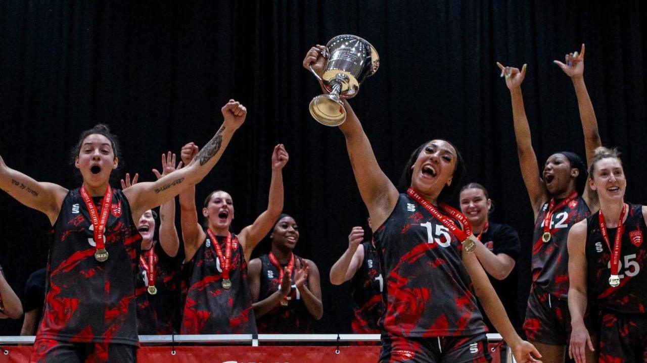 A woman holds a large silver trophy in the air while cheering. Seven other members of the team, who are all wearing lack and red sports vests, are behind her. They are cheering with their arms in the air in celebration.