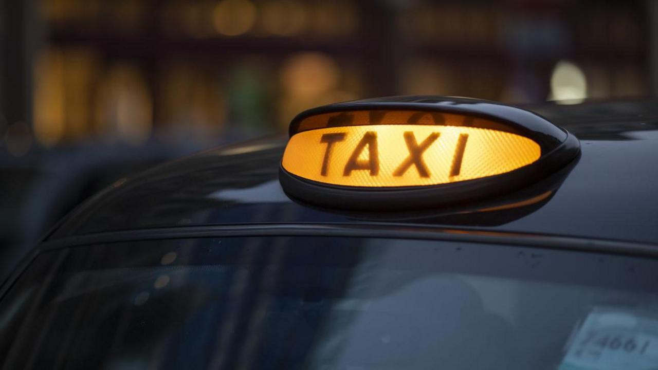 A close-up shot of a lit-up yellow taxi sign on the top of a black cab. It says "TAXI" in bold across it. The background is blurred.