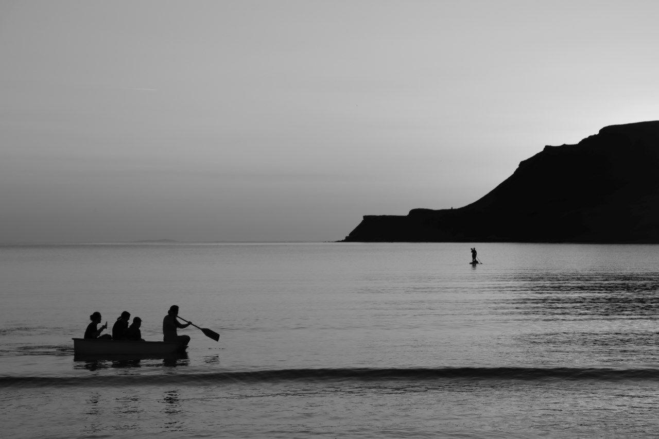 A canoe carrying four people seen in silhouette on a river. A person on a paddleboard is seen in the distance.