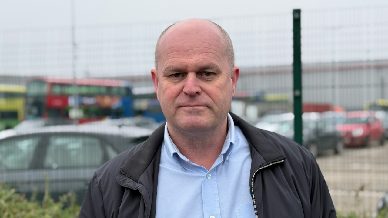 Dave Telford has a shaved head. He is standing outside a bus depot.