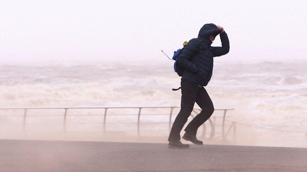 A person walks along a promenade with a raging sea in the background, they're holding onto their hood as sea sprays up due to high winds, taken in Blackpool on 7 December.