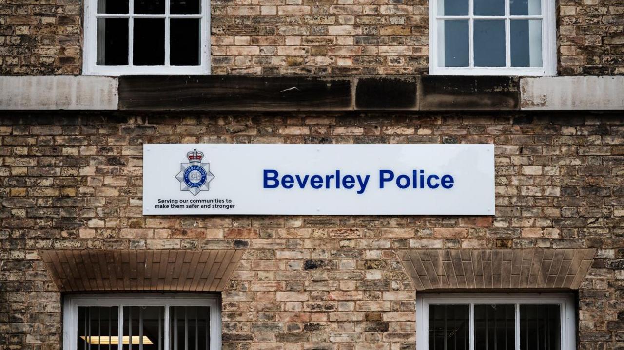 A sign which reads 'Beverley Police' with a white background positioned on a sandy brick walled building with four windows. A police logo can be seen to the left of the sign.