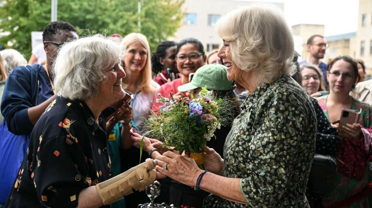 Queen Camilla leaves Royal United Hospitals Bath NHS Foundation Trust. She is smiling as she accepts a small posy of flowers from a woman with short grey hair. The woman has a wrist support on and the Queen is touching her hand. Behind them there are several people watching on and taking photos on their phones.