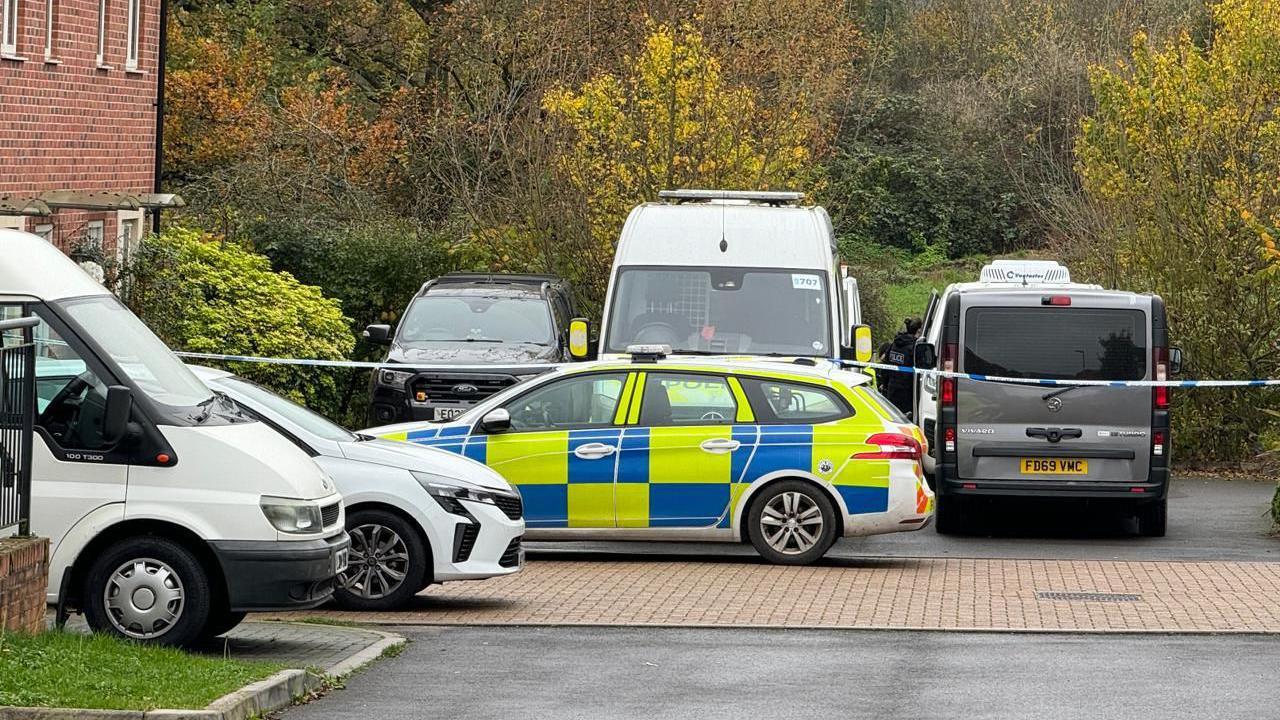 A marked police car parked next to five other vehicles. Police tape can be seen across the end of a quiet cul-de-sac. Beyond the end of the road is a field and some trees. 