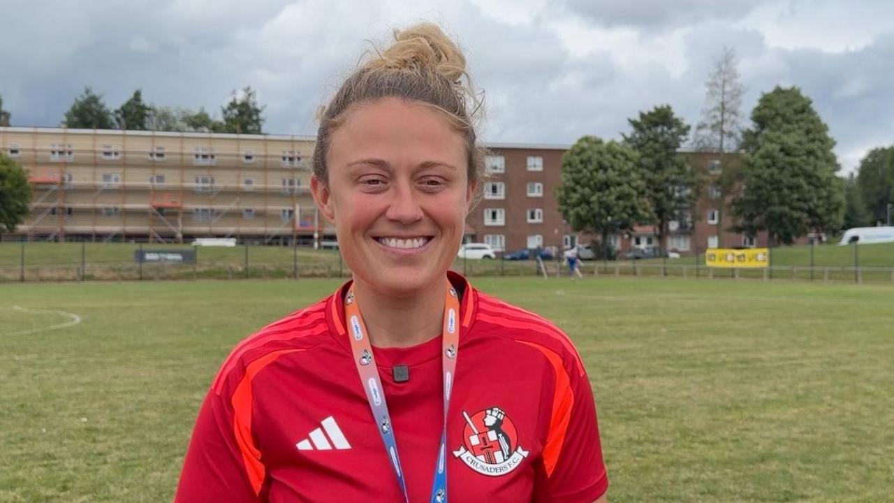 Heather Mearns standing on a football pitch wearing a red top with the Crusaders logo on it. She has a blue and orange lanyard around her neck and blonde hair pulled up into a bun on top of her head.