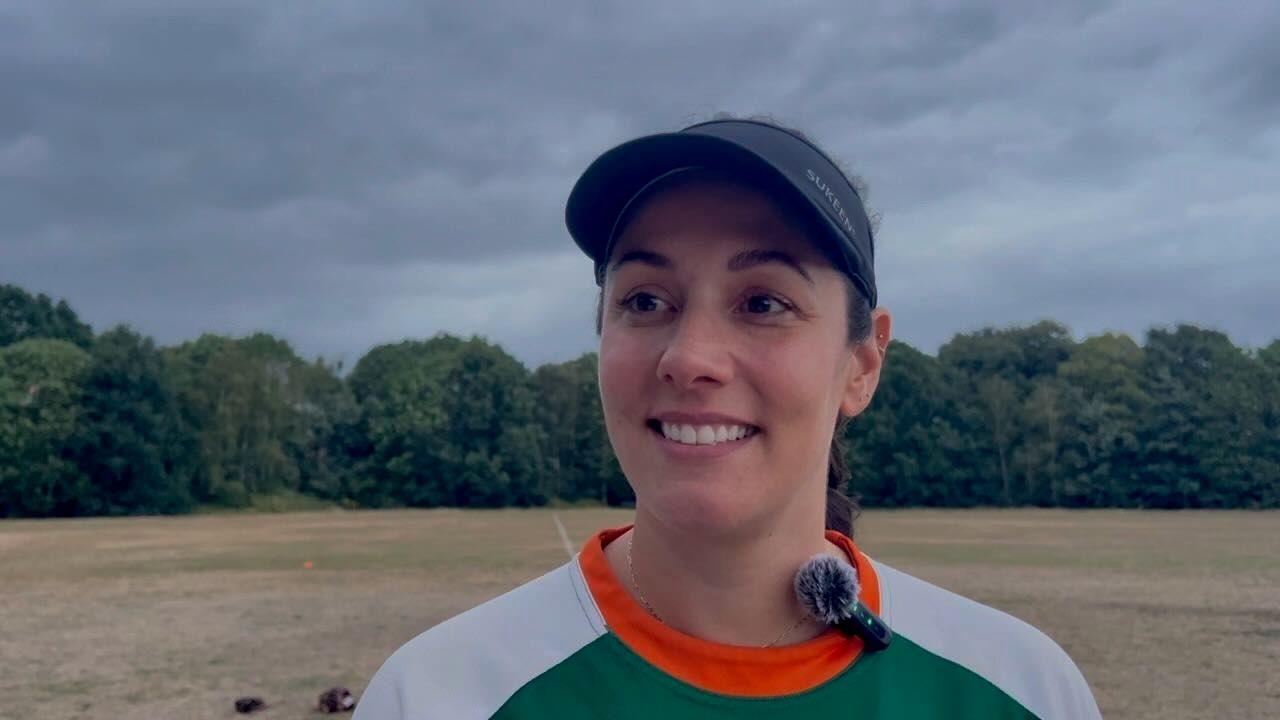 Dom Kleeman has dark hair tied back in a ponytail and is wearing a black sports cap. She's wearing an orange, green and white softball top and is standing on Wandsworth Common (a dry grassy field with trees in the background).