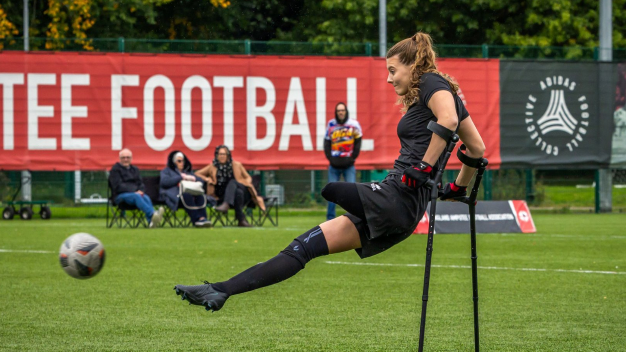 A woman with an amputated leg kicks a ball - she is leaning on her crutches while her body is swinging upwards into the air. She wears a football kit comprised of a black t-shirt, black shorts, and a long black sock and trainer on one leg. She is on a football pitch and three people in chairs and one person standing up watch her in the background.