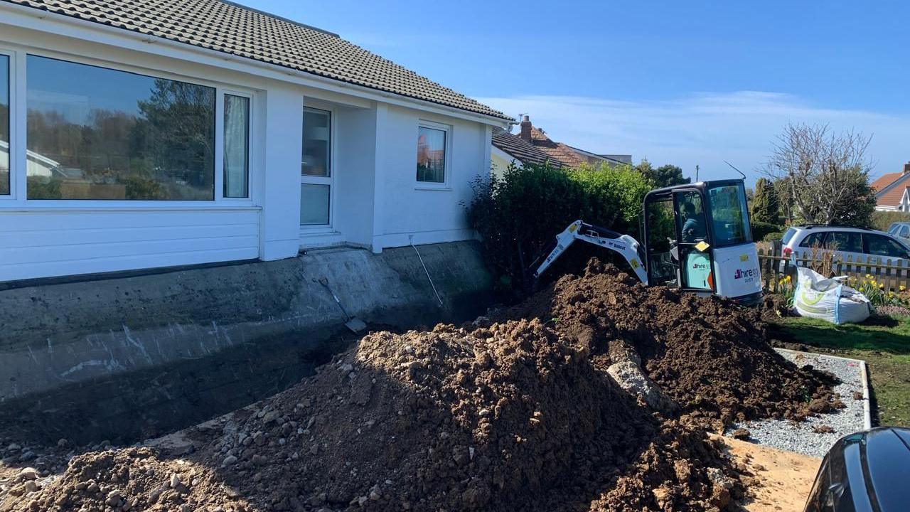 A man sits in a white digger and works to remove earth from underneath a house in Guernsey on a sunny day.