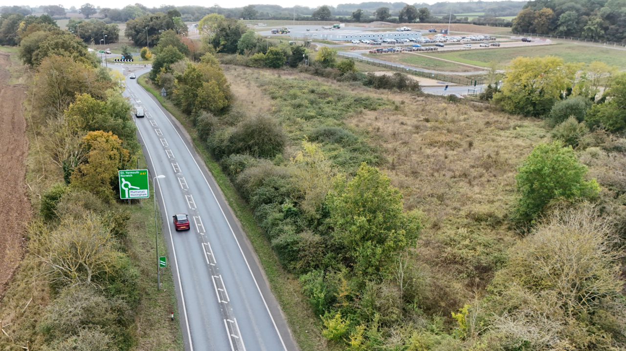 A road is going up towards a roundabout on the left hand side of the picture. Trees surround either sides of the roads, and 2 cars are driving towards the roundabout. 