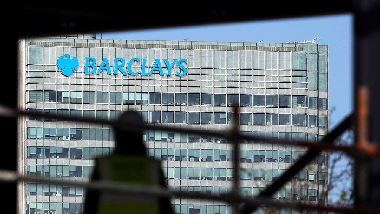 The Barclays logo is seen on the side of the Barclays building in Canary Wharf, with a silhouetted construction worker out of focus in the foreground.