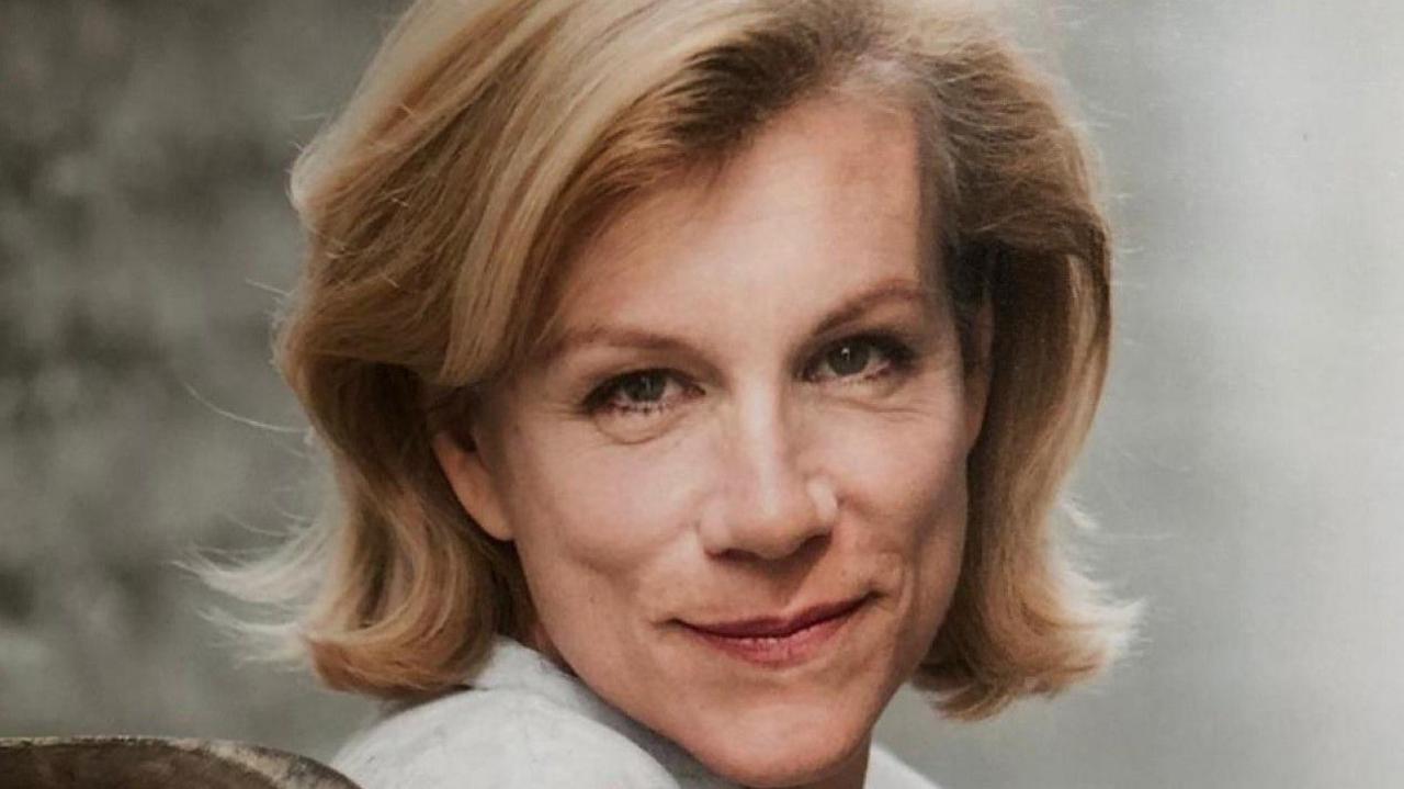 A close-up photo of a woman in front of a plain grey background. She has blonde hair and is wearing a cream top.