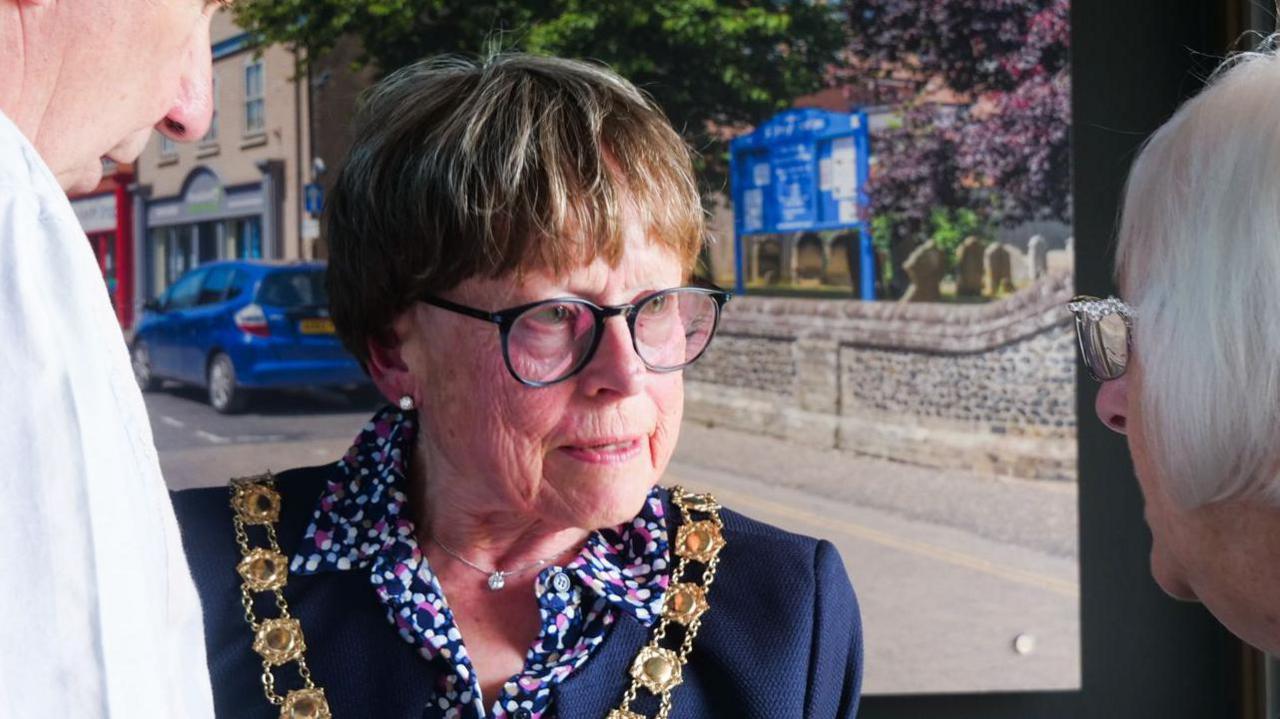 Victoria Holliday is wearing a gold ceremonial chain over a navy blue jacket. Beneath that, she has a patterned blouse. She is talking to a man and a woman.