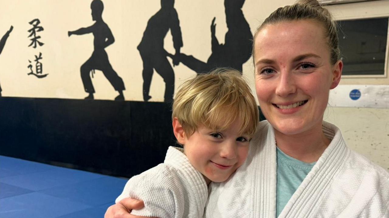 A woman and a child smiling at the camera. They are both wearing a white judo jackets. The woman, on the right, has blonde hair which has been tied up into a bun. The boy, on the left, has a short mop of blonde hair. The floor is covered in blue judo maps. There are large silhouettes of people in judo poses on the wall.