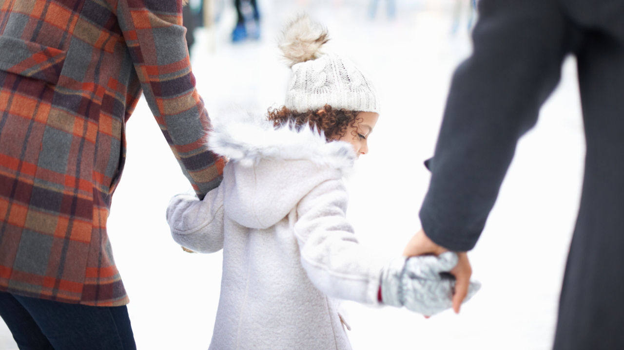 A young girl in a white coat and hat holding on to the hands of a man and a woman on an ice rink