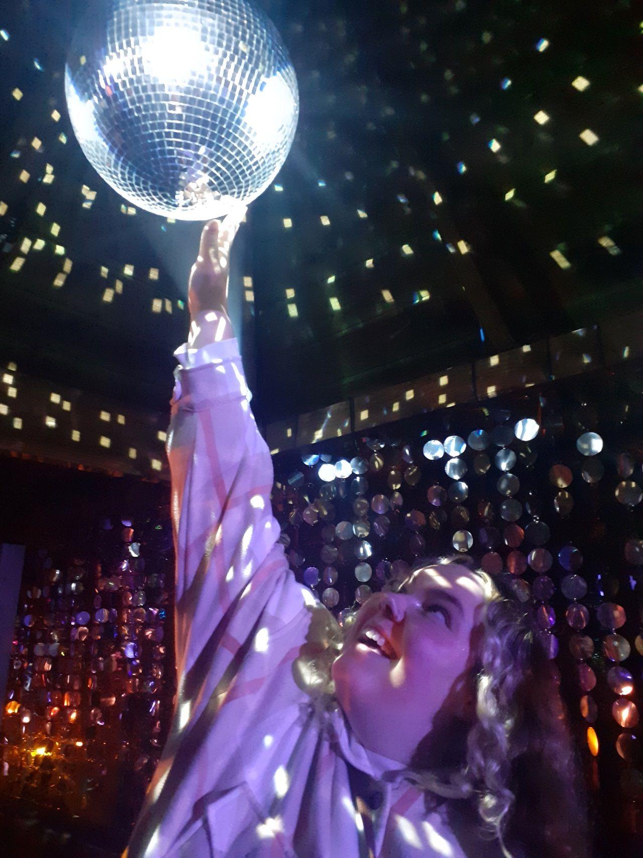 A young girl holds her hand up to a disco ball surrounded by coloured lights.