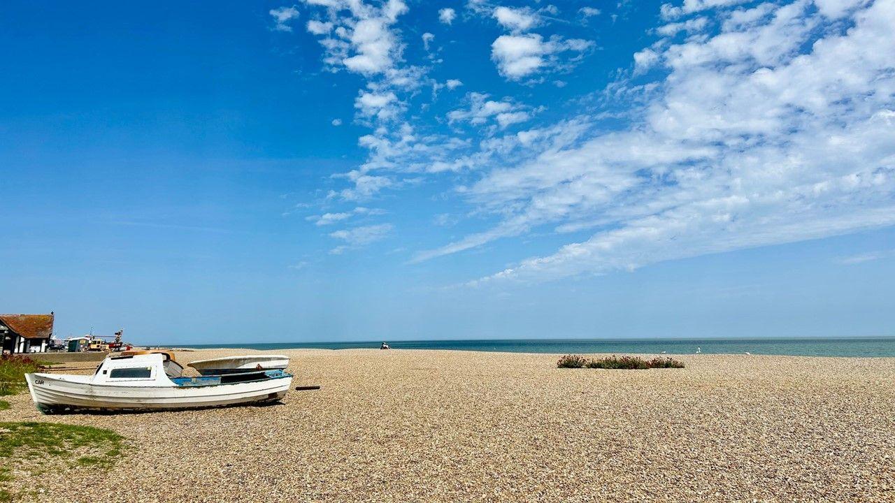 A wide view of a flat beach with a boat on the right, the sea in the distance and a blue sky with streaks of white cloud