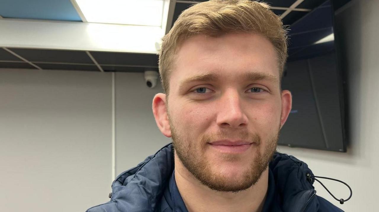 Will is in an office with a screen in the background. He is wearing his navy Cardiff Rugby training kit and coat. He has short, fair hair and a beard.