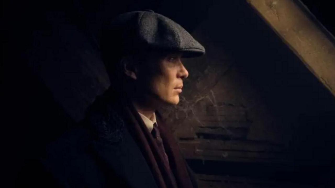 A man with a flat cap standing in the dark with only his face illuminated.