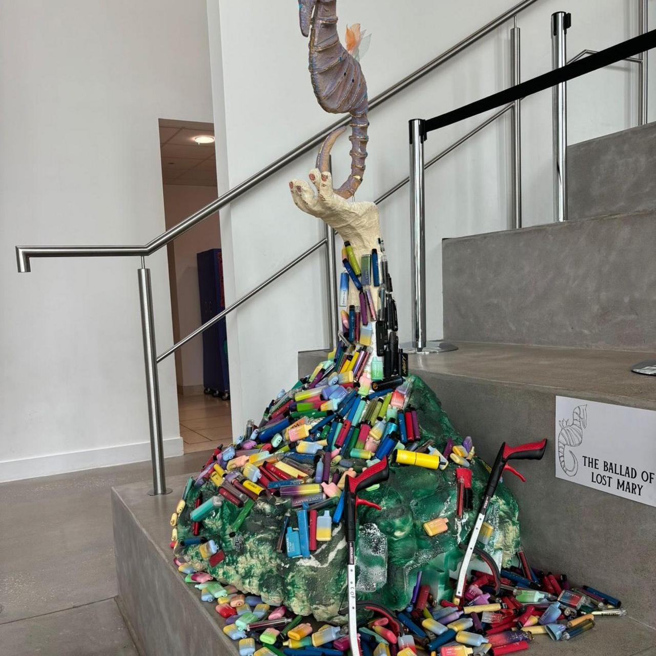 A sculpture designed using 4,500 disposable vapes collected by volunteers. The sculpture also has a grey seahorse on top and is positioned at the bottom of a flight of stairs.