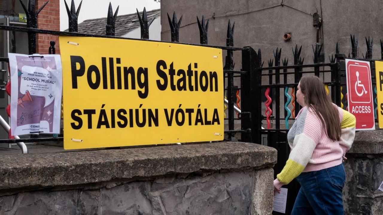 The exterior of a polling station in Dublin. There is a large yellow sign ziptied to the black iron fence outside the building. A young woman with long, brunette hair, a pink, grey, yellow and white cardigan and dark wash blue jeans is walking through the front gate.