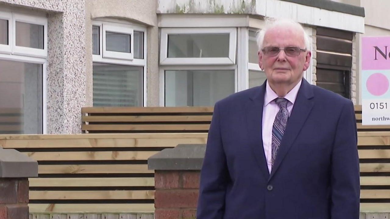 Terence Crolley, wearing a navy blue suit over a light pink shirt, stands on a street with terraced houses in the background.