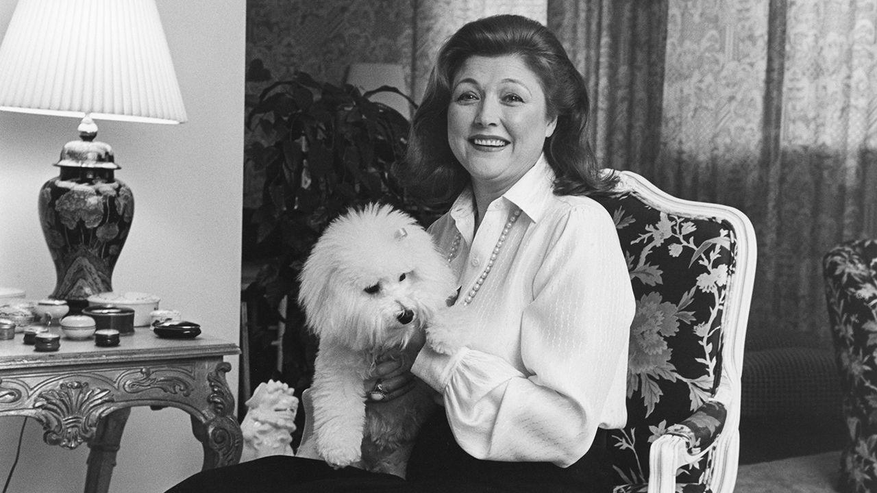 Barbara Taylor Bradford sits in a chair holding a fluffy dog and smiling next to a table with a lamp and trinkets on top, black and white photo taken circa 1980