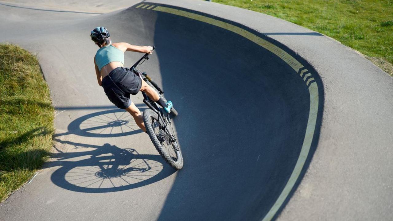 A female cyclist seen from behind riding a mountain bike round a bend in a steeply curved track. She is standing up on her pedals and wearing navy shorts, a pale blue crop top and helmet and her hair is tied back in a short ponytail. The sun casts her shadow to her left and grass can be seen adjacent to the track.