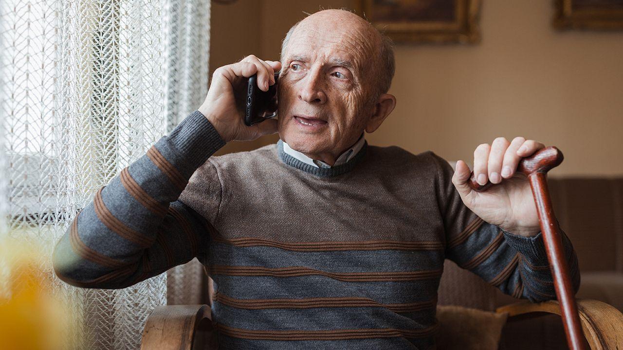 An elderly man holds a phone to his ear as he makes a call while looking out the window at home. He's sat on a chair near the window, which is covered by light white transparent curtains, and wears a jumper over a shirt, and holds a walking stick in his free hand.