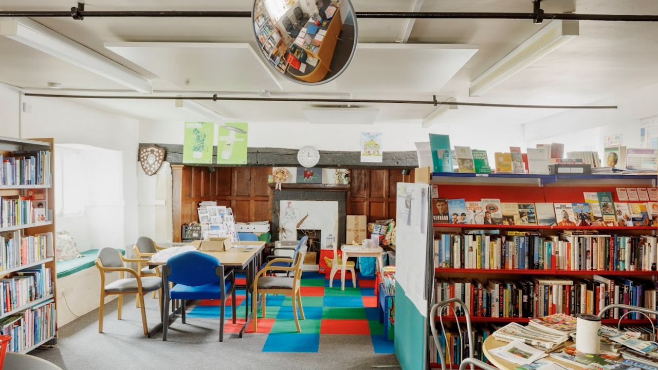 A library filled with books on book cases in a low ceiling room. There are tables and chairs in the middle of the room on a multi-coloured checked carpet. There's a beam and a fireplace at the back of the room.