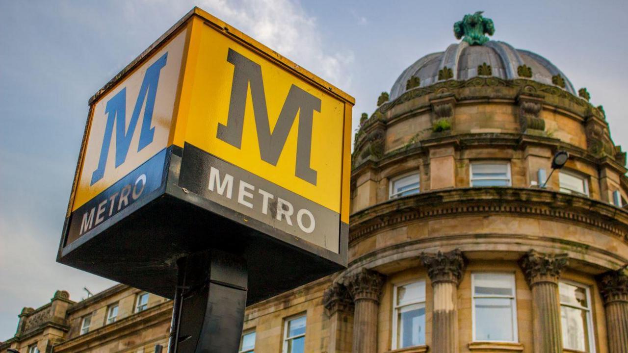 A sign for the Tyne and Wear Metro. The yellow and black sign is a large cube on top of a pole. It displays the word Metro and it's M logo. A building from the Grainger Town area of Newcastle city centre stands in the background.